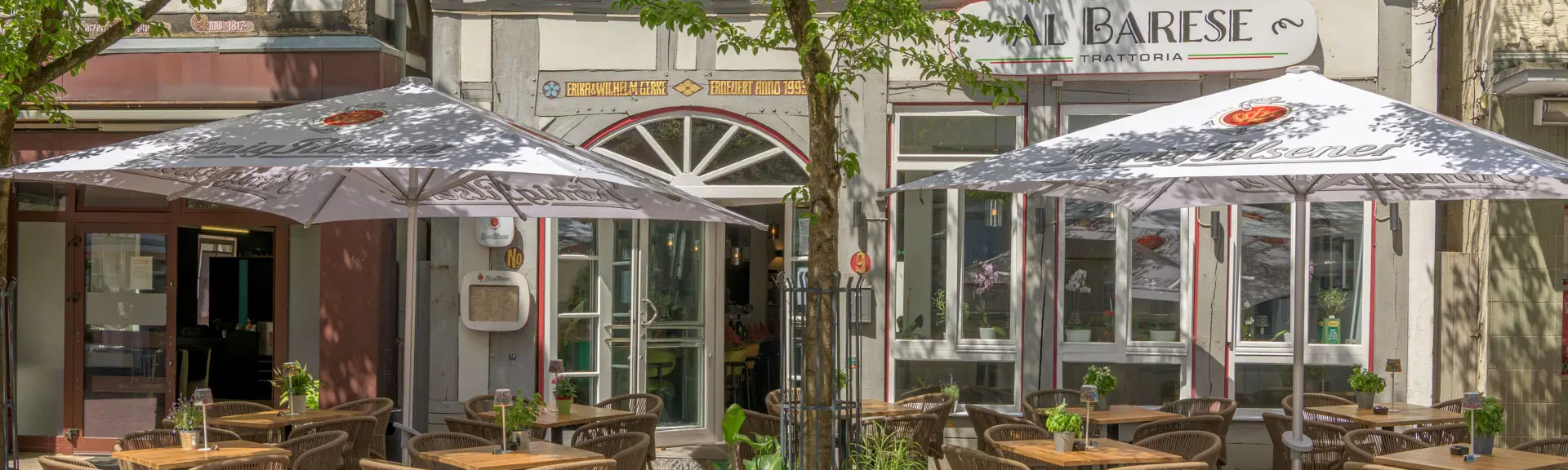 Außenterrasse eines Restaurants mit Tischen, Stühlen und zwei großen Sonnenschirmen vor einem Gebäude mit Bogenfenster und Schriftzug 'AL BARESE'.
