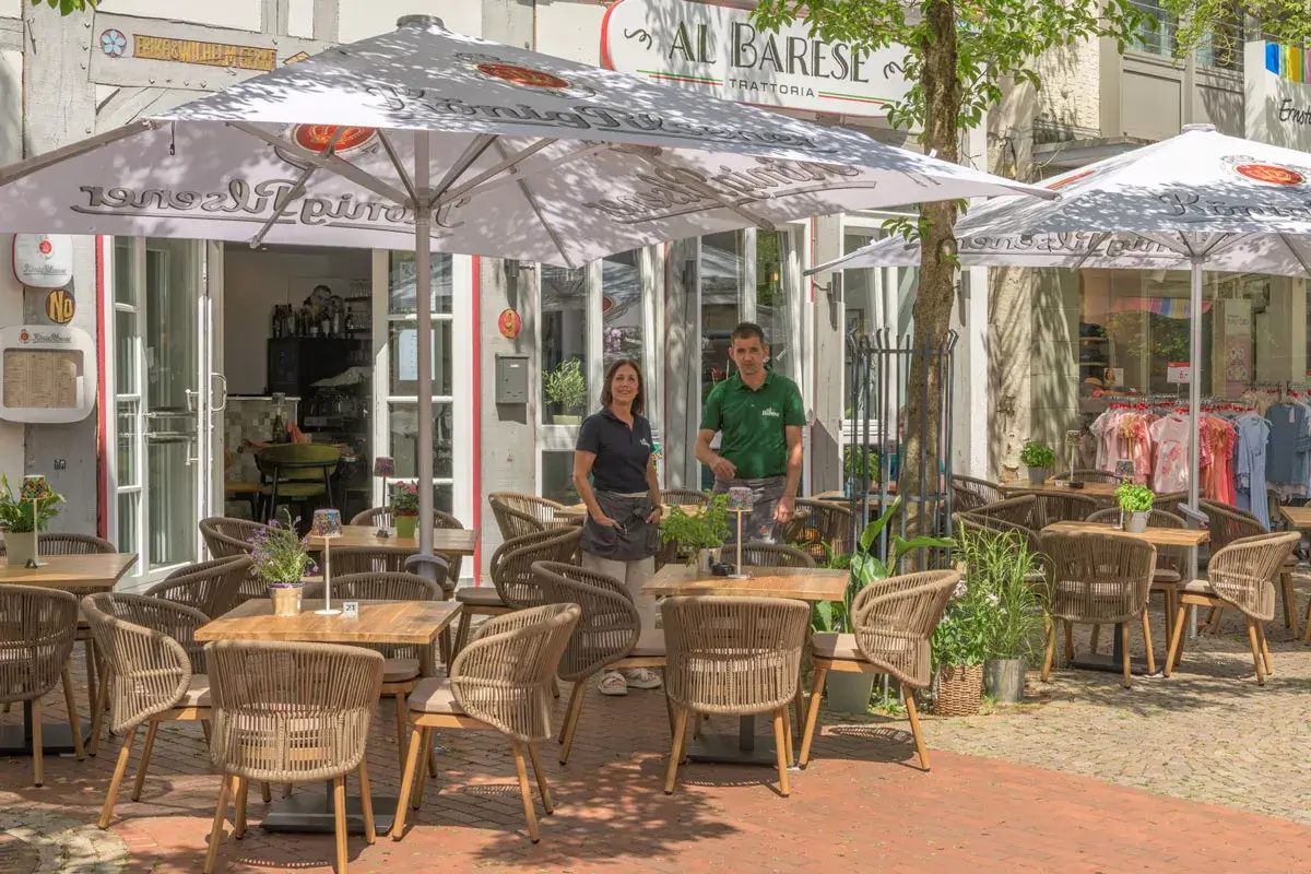 Kellnerin und Kellner stehen vor einem Restaurant mit Tischen, Stühlen und zwei großen Sonnenschirmen vor einem Gebäude mit Bogenfenster und Schriftzug 'AL BARESE'.
