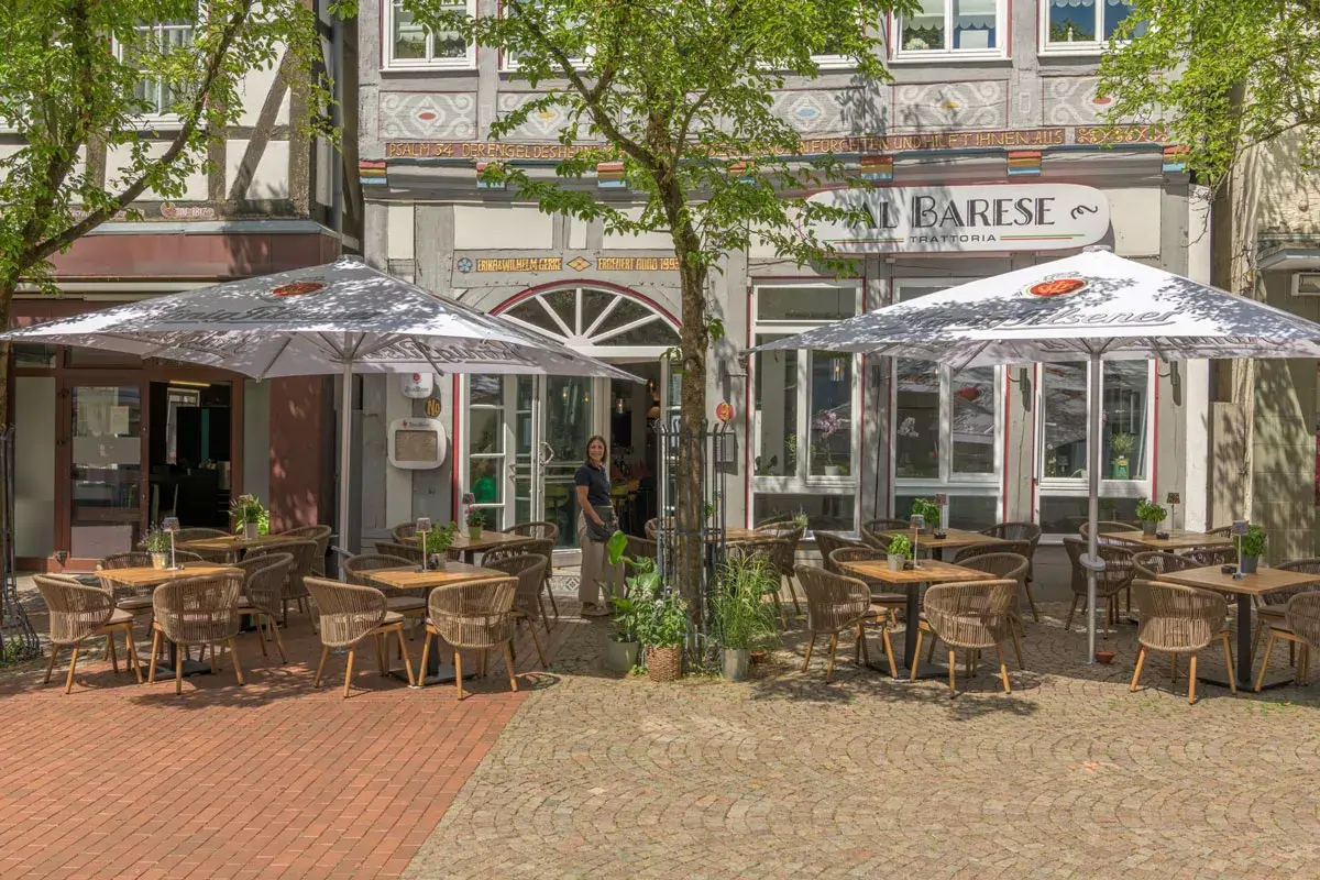 Kellnerin steht auf der Außenterrasse eines Restaurants mit Tischen, Stühlen und zwei großen Sonnenschirmen vor einem Gebäude mit Bogenfenster und Schriftzug 'AL BARESE'.