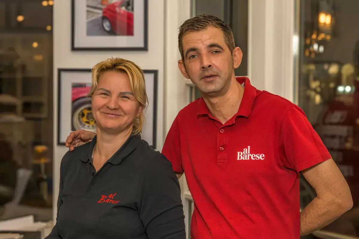 Zwei Personen in schwarzen und roten Poloshirts mit dem Schriftzug 'al Barese', stehend in einem Raum mit Bildern an der Wand.