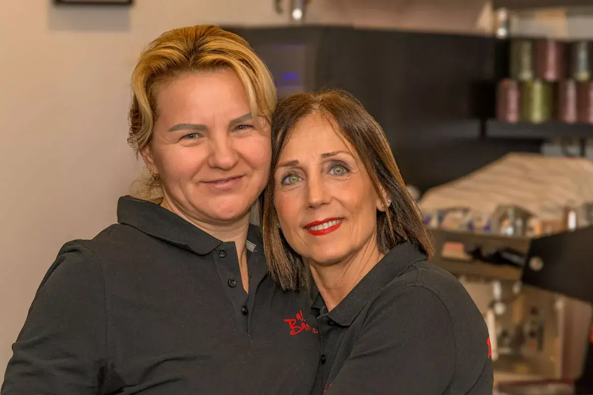 Zwei Frauen in schwarzen Poloshirts mit rotem Logo stehen in einer Küche oder Cafeteria, im Hintergrund Kaffeemaschine und Regale mit Bechern.