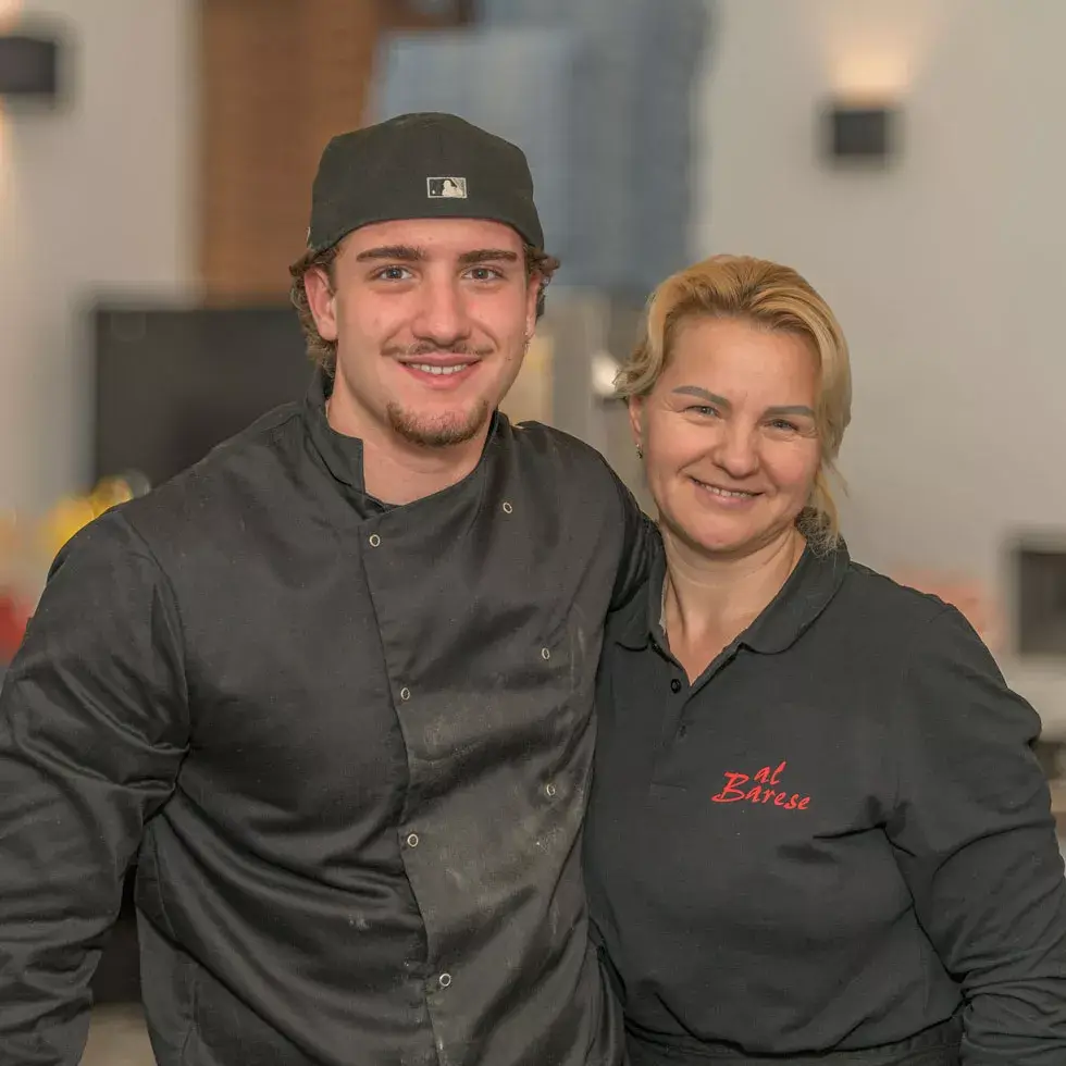 Zwei Personen in Arbeitskleidung, einer trägt schwarze Kochjacke und Mütze, die andere schwarzes Poloshirt mit rotem Schriftzug 'al Barese'.