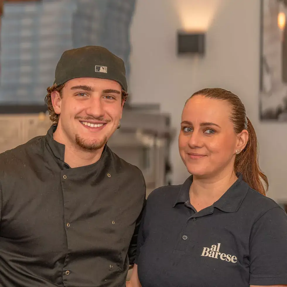 Zwei junge Personen in Arbeitskleidung, einer trägt schwarze Kochjacke und Mütze, die andere schwarzes Poloshirt mit weißem Schriftzug 'al Barese'.