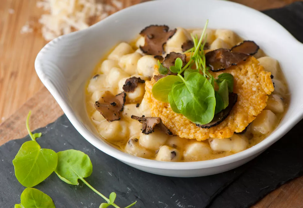 Weiße Schüssel mit Gnocchi in cremiger Sauce, garniert mit schwarzen Trüffelscheiben, knusprigem Käsechip und grünen Blättern auf Holz- und Schieferuntergrund.