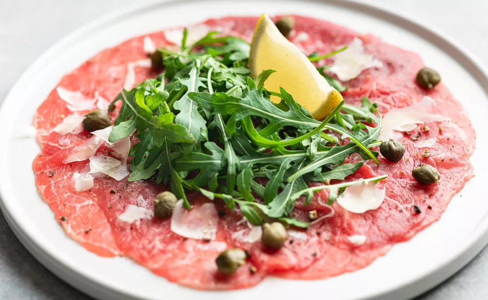 Rinder-Carpaccio auf weißem Teller mit Rucola, Kapern, Parmesanspänen und Zitronenspalte