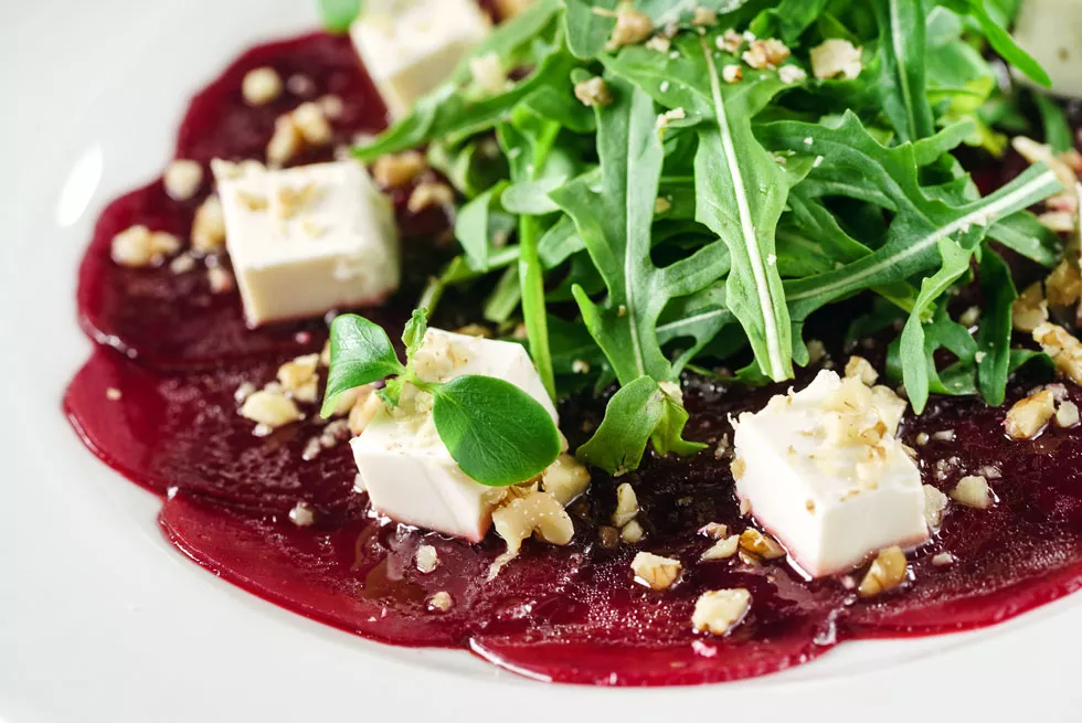 Rote-Bete-Scheiben mit Rucola, Feta und gehackten Walnüssen auf weißem Teller angerichtet.