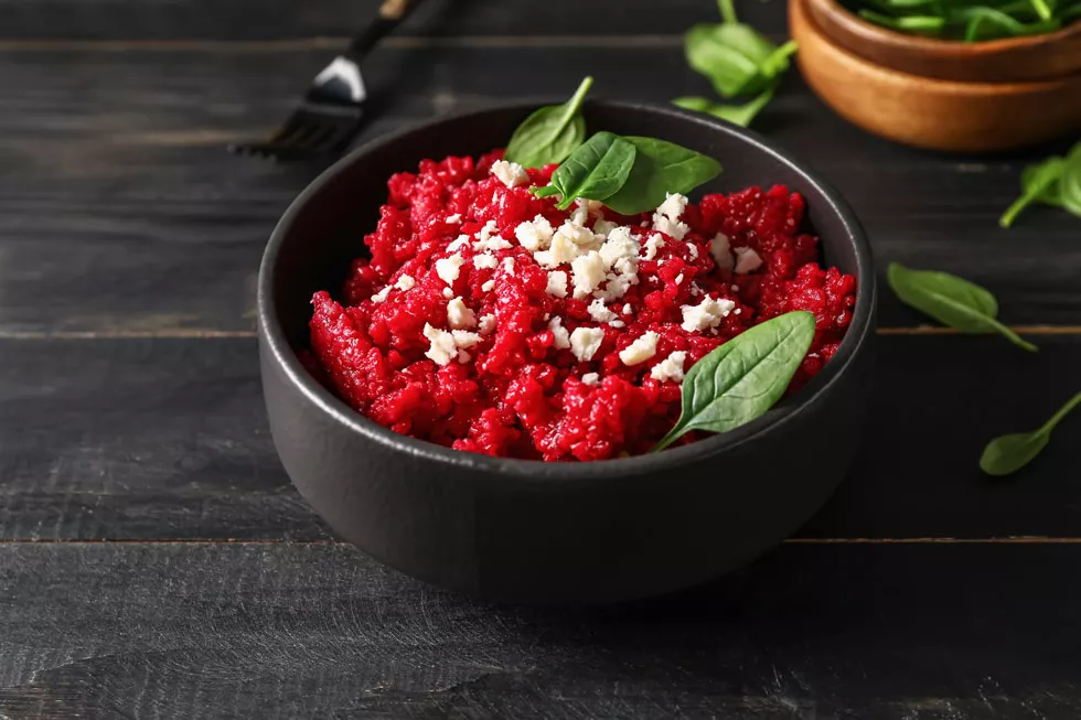 Schwarze Schale mit rotem Risotto und Rote Beete Stückchen, garniert mit zerbröseltem weißen Käse und frischen Basilikumblättern auf dunklem Holztisch.