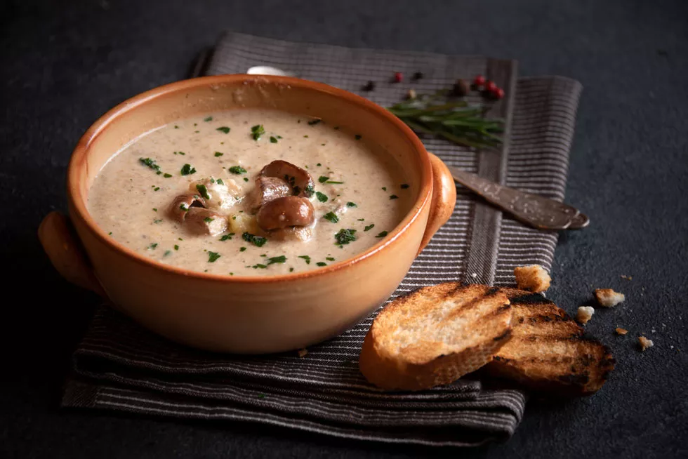 Braune Keramikschale mit cremiger Pilzsuppe, garniert mit Petersilie, daneben zwei geröstete Brotscheiben auf dunkelgrauem Stoff.