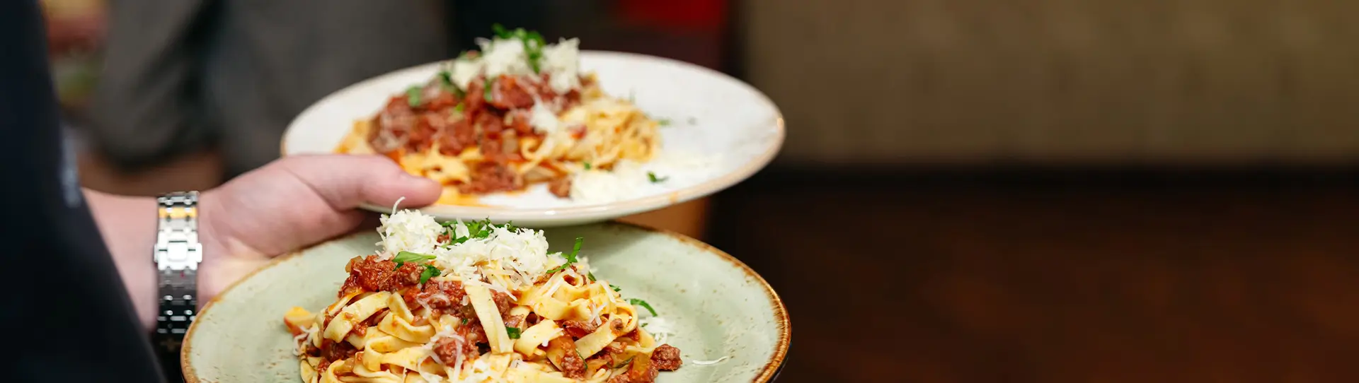 Zwei Teller mit Pasta, jeweils mit Hackfleischsauce und geriebenem Käse, werden von einer Person serviert.