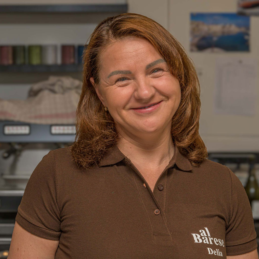 Frau mit schulterlangen, gewellten Haaren trägt ein braunes Poloshirt mit weißem Schriftzug 'al Barese' im Innenraum mit Regalen und Bildern im Hintergrund