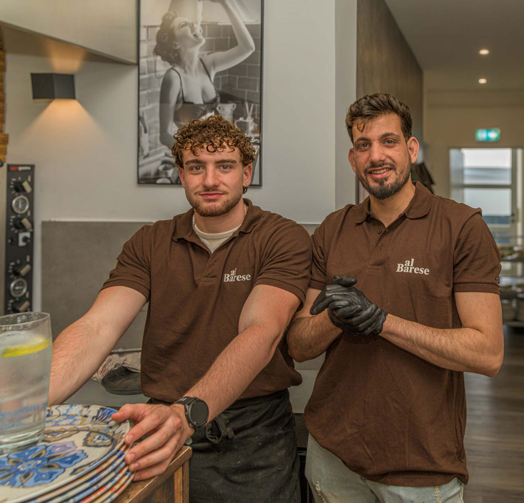 Zwei Männer in braunen Poloshirts mit dem Schriftzug 'al Barese' stehen hinter einem Tresen mit einem Stapel bunter Teller und einem Glas Wasser mit Zitronenscheibe