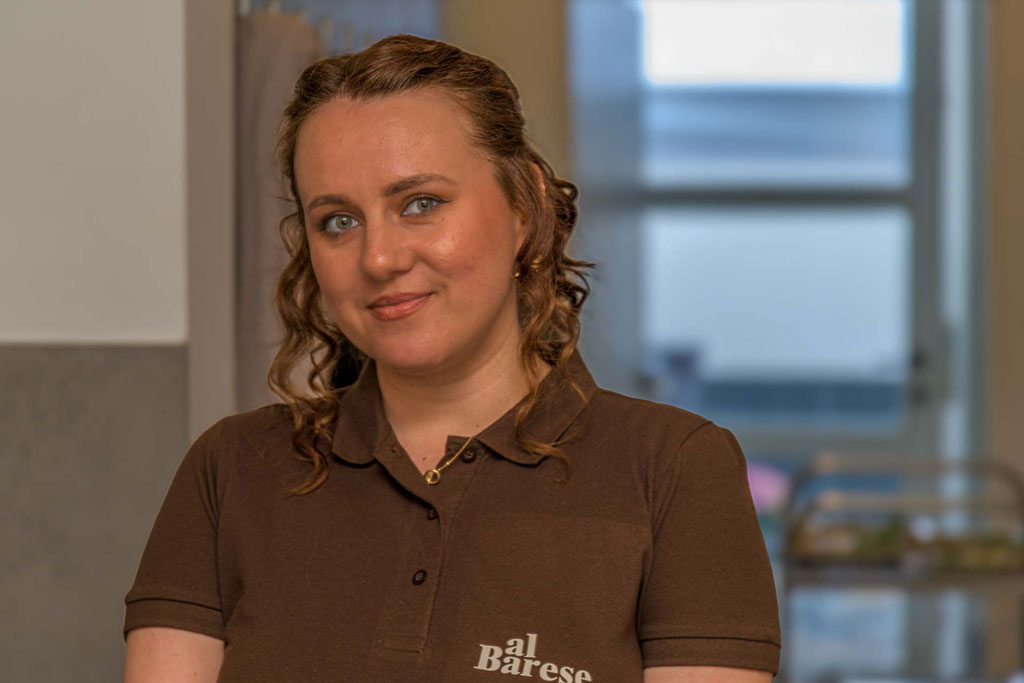 Frau mit lockigem Haar trägt ein braunes Poloshirt mit dem Schriftzug 'al Barese' vor einem Fenster mit Blick auf eine Stadt.
