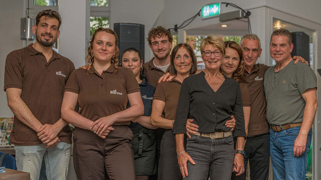 Gruppe von sieben Personen steht eng beieinander in einem Innenraum, sechs tragen braune Poloshirts, eine Person trägt schwarze Bluse