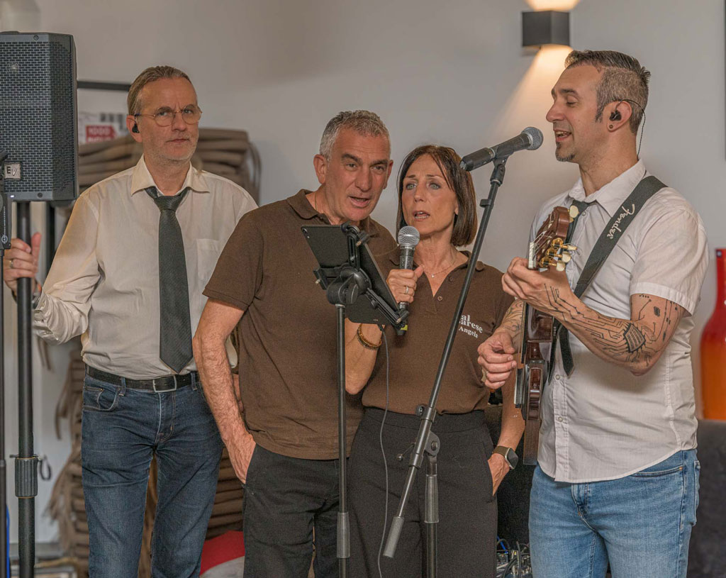 Vier Personen stehen mit Mikrofonen und einem E-Gitarrenverstärker in einem Raum, eine Person spielt Gitarre, zwei halten Mikrofone, eine hält einen Lautsprecherständer