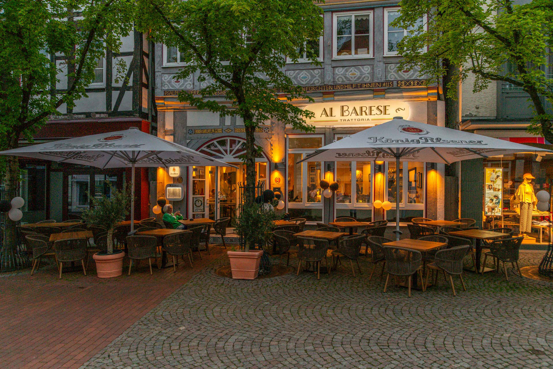 Außenbereich eines Restaurants mit Tischen, Stühlen, großen Sonnenschirmen und Bäumen vor einem Fachwerkgebäude mit beleuchtetem Schriftzug