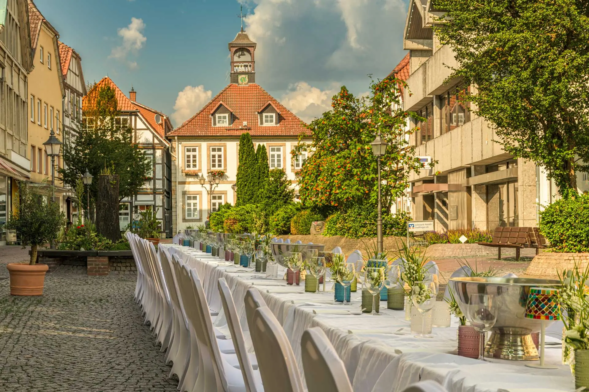 Langer festlich gedeckter Tisch mit weißen Stühlen in einer gepflasterten Altstadtstraße vor historischen Fachwerkhäusern und einem Gebäude mit Uhrturm