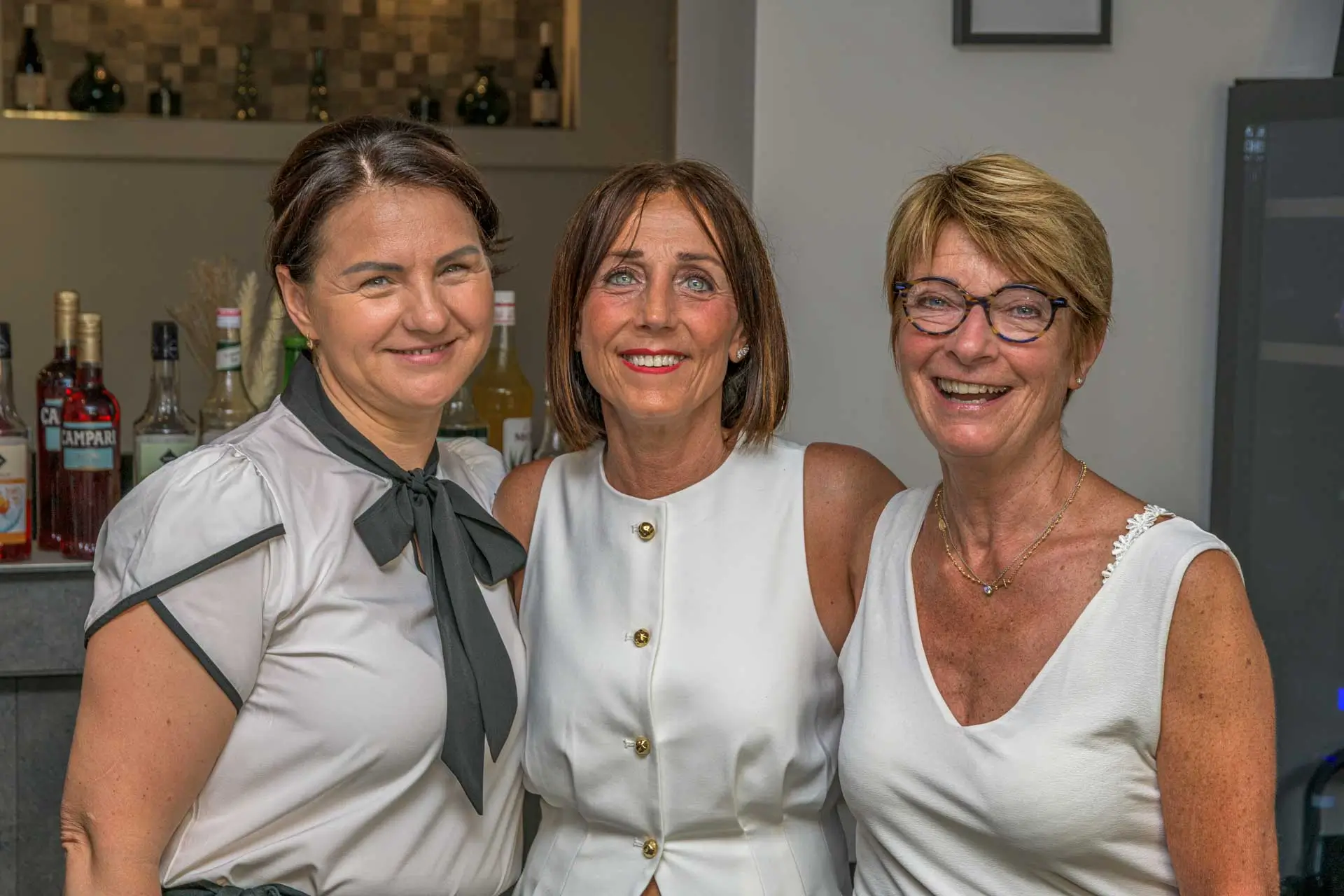 Drei lächelnde Frauen stehen eng beieinander in einem Innenraum mit einer Bar im Hintergrund