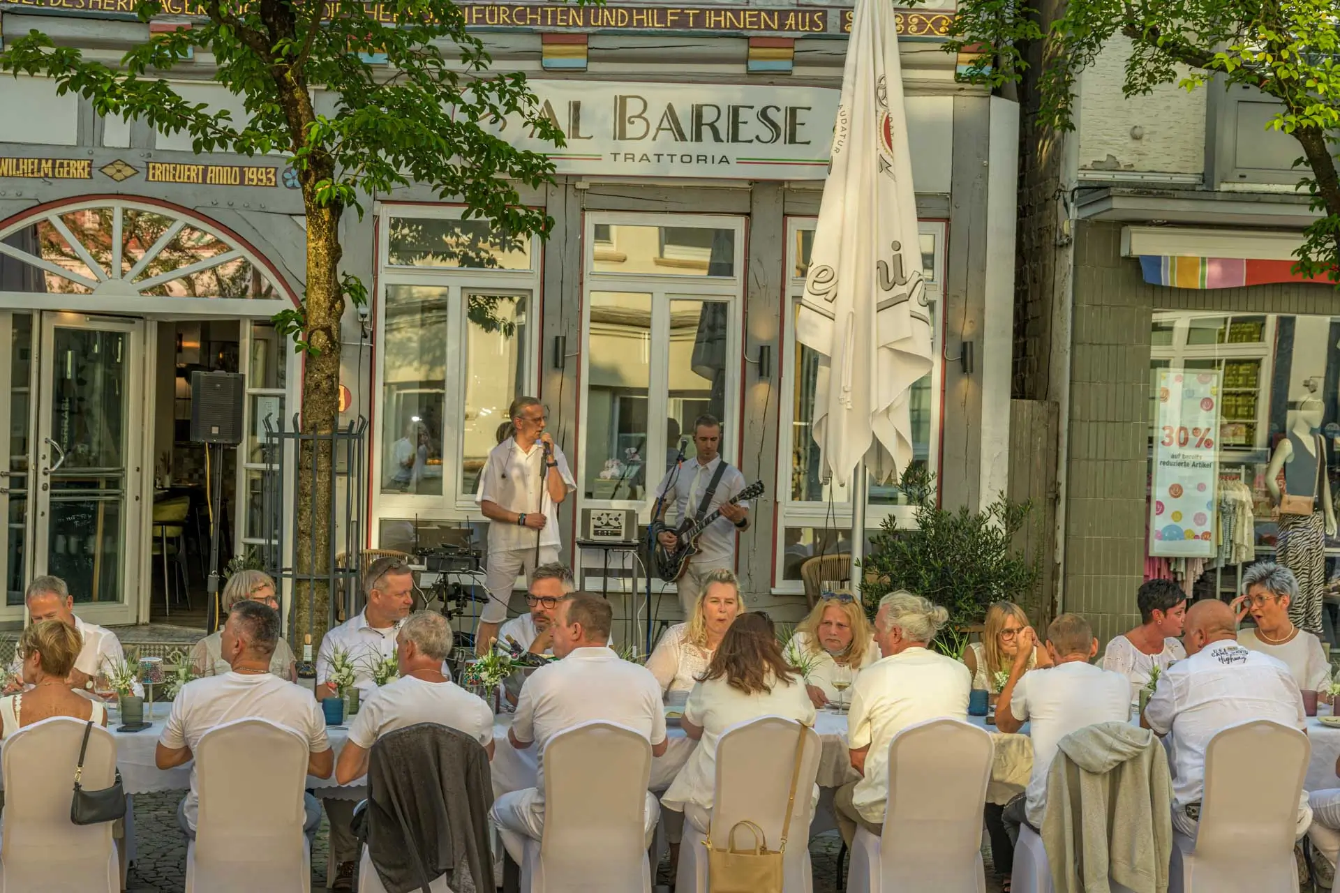 Menschen sitzen an einem langen Tisch im Freien vor einem Restaurant mit der Aufschrift 'AL BARESE', während zwei Musiker mit Mikrofon und Gitarre spielen