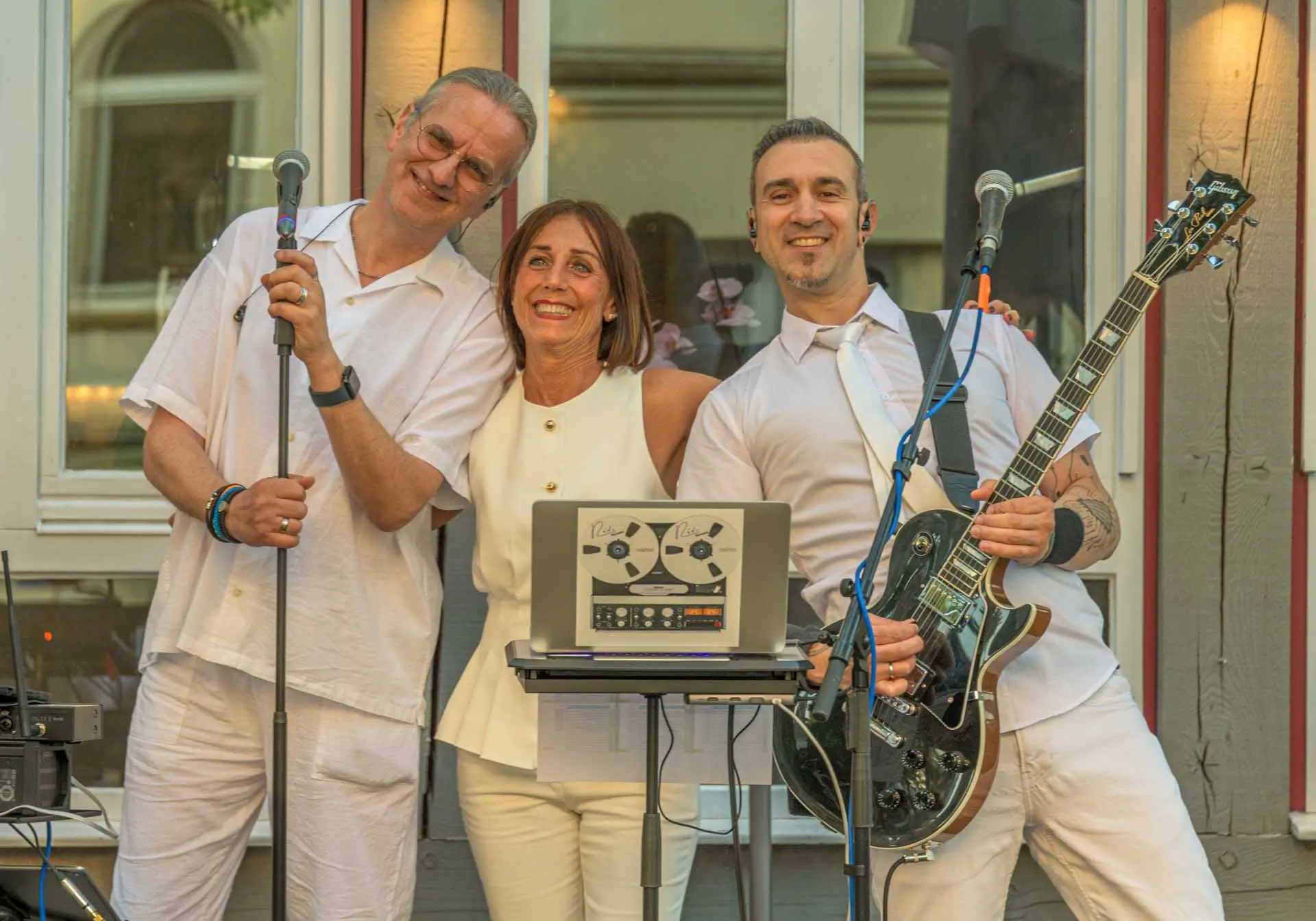 Drei Personen in weißer Kleidung posieren vor einem Gebäude, zwei Männer halten Mikrofon und E-Gitarre, eine Frau steht in der Mitte lächelnd