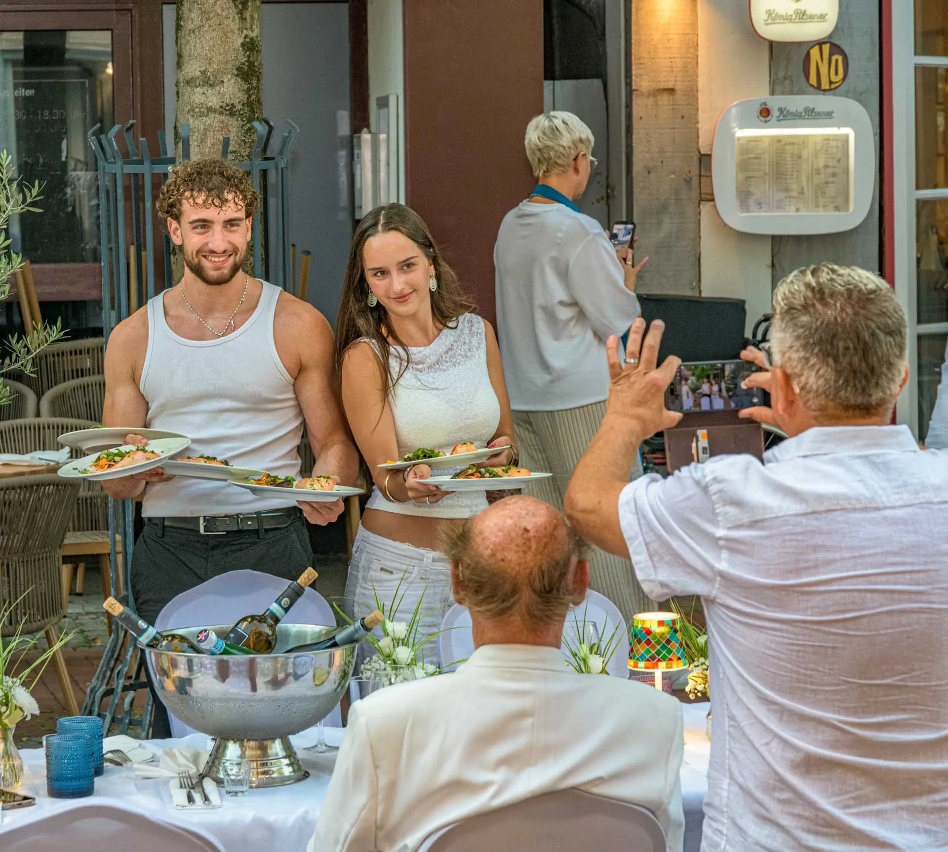 Zwei junge Bedienungen mit Tellern posieren vor einem Tisch, an dem zwei Männer sitzen, während ein weiterer Mann ein Foto macht