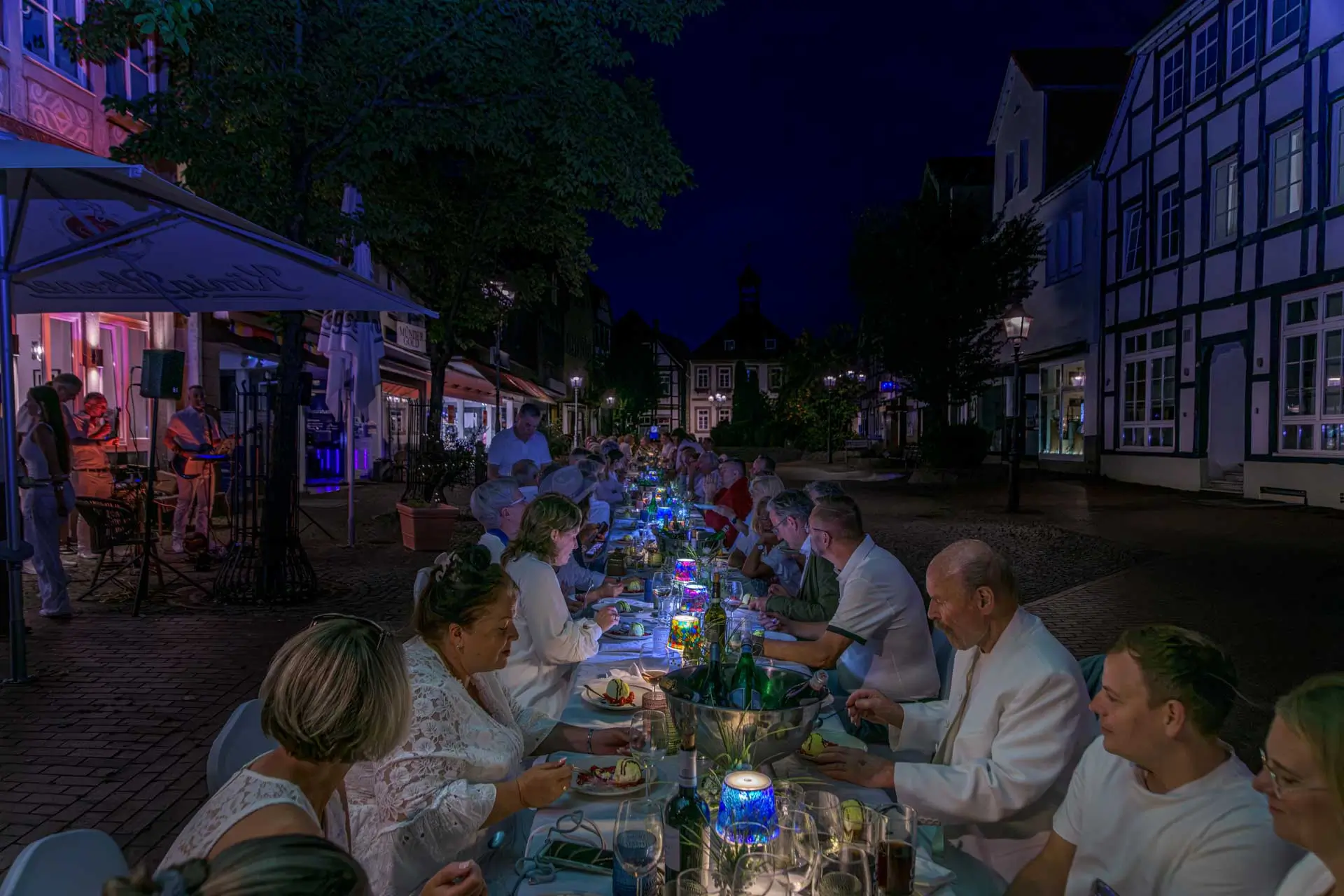 Lange Tafel mit Menschen in weißer Kleidung bei Nacht in einer beleuchteten Straße mit Fachwerkhäusern